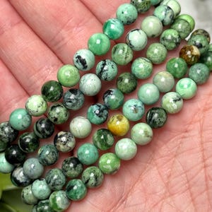May include: Strands of round, green gemstone beads in varying shades, including light, medium, and dark greens, with some beads featuring black and yellow inclusions. The beads are smooth and appear to be strung together, suitable for jewelry making.