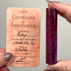 May include: A deep red ruby crystal wand with dark inclusions is held next to a certificate. The certificate reads "Certificate of Resonance" and lists the crystal name as "Ruby". The frequency is listed as 12695 H2, G, tested on 2/27/26.