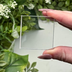 May include: A clear, square glass piece with a thin, silver-colored border. The glass is held up against a backdrop of green foliage and white flowers. The glass appears to be a small, transparent square.