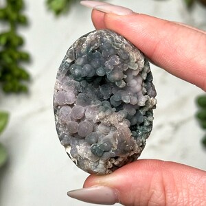 May include: A smooth, oval-shaped agate stone with a unique pattern of purple, green, and white. The stone has a textured surface with small, round bumps.