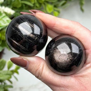 May include: Two polished black obsidian spheres with reflective surfaces. The stones feature natural inclusions and subtle metallic sheen patterns visible across their smooth, rounded forms. These dark, circular mineral specimens are held together.