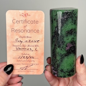 May include: A cylindrical ruby in zoisite crystal with green and black mottled patterns, accompanied by a peach-colored Certificate of Resonance card. The card displays the text: Certificate of Resonance, Crystal Name Ruby in Zoisite, Frequency (Hz) / Musical Note 33477Hz, C, Tested on 7/22/26, and Loving Thyself Rocks.
