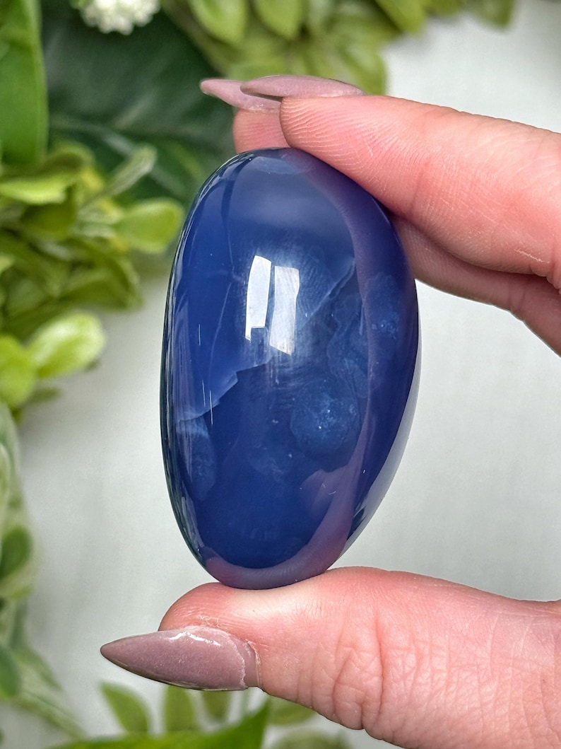May include: A smooth, oval-shaped, polished stone in a deep blue hue. The stone has subtle internal patterns and a glossy surface, reflecting light. The object is held between fingers, with green foliage in the background.