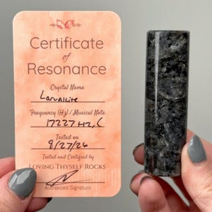 Pode incluir: Uma torre de cristal Larvakite cilíndrica com uma aparência cinzenta e preta salpicada, acompanhada por um cartão de Certificado de Ressonância cor de pêssego com o texto Crystal Name Larvakite, Frequency 17227Hz, C, e Loving Thyself Rocks.