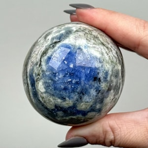 May include: A polished, spherical stone featuring a deep blue center surrounded by swirling patterns of white, gray, and black mineral inclusions. The surface is smooth and reflective, showcasing a natural crystalline texture within the orb.
