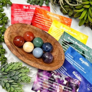 Könnte beinhalten: Sieben glatte, runde, polierte Edelsteine in einer Holzschale. Die Steine sind rot, orange, gelb, grün, blau und lila. Jeder Stein ist mit seinem entsprechenden Chakra und seinen heilenden Eigenschaften beschriftet.