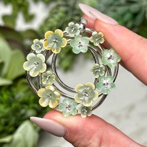 Peut inclure: Pendentif argenté avec un motif floral. Le pendentif présente un cadre circulaire orné de petites fleurs tridimensionnelles dans des tons de jaune pâle et de vert. Les fleurs sont disposées autour du cadre, créant une esthétique délicate et féminine.