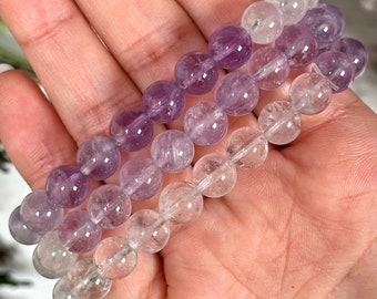 May include: Two bracelets made of clear and purple beads. The bracelets are layered on top of each other and are being held in a hand.
