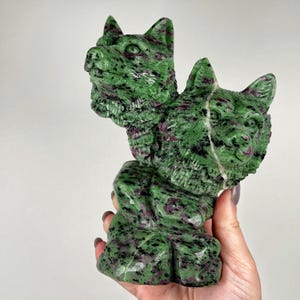 May include: A carved stone sculpture of two wolf heads, one above the other. The sculpture is green with dark red and black inclusions. The wolf heads have detailed features and are set on a textured base.