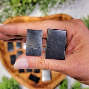 Peut inclure: Deux pierres rectangulaires polies, l'une grise foncée veinée de clair, l'autre noire, sont tenues dans une main. D'autres pierres similaires sont visibles dans un bol en bois en arrière-plan. Les pierres sont probablement destinées à la bijouterie ou à l'artisanat.