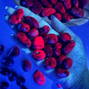 May include: A hand holding a collection of red tumbled stones. The stones are smooth and polished, and they have a deep red color. The stones are illuminated by a blue light, which makes them appear to glow.