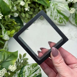 Op de afbeelding: Een zwarte vierkante displaykoffer met een helder glazen venster, vastgehouden door een hand met verzorgde nagels. De koffer is omgeven door groene bladeren en witte bloemen, wat een natuurlijke achtergrond creëert. De koffer is ontworpen om kleine items te presenteren.