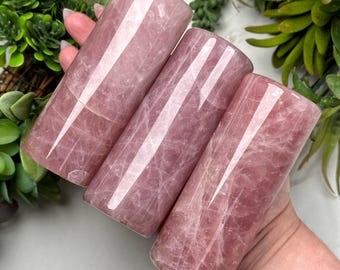Puede incluir: Tres cilindros pulidos de cuarzo rosa. Las piedras rosas tienen una calidad translúcida con vetas internas visibles. Los cilindros se sostienen en una mano, con plantas verdes en el fondo. Los cilindros son lisos y redondeados.