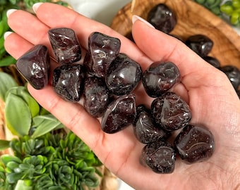 May include: A collection of tumbled red garnet stones in a hand. The stones are smooth and polished, with a deep red color.