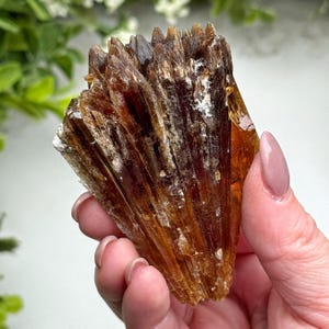 May include: A cluster of brown and amber crystal formations. The crystals have a layered, textured appearance with sharp, pointed tips. The mineral is held in a hand, with a blurred green background.