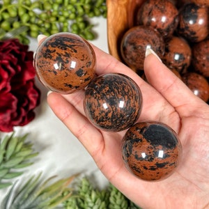 May include: Three polished red and black jasper spheres, each with a unique pattern of black and red markings. The spheres are displayed on a hand.