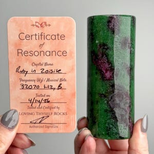 Peut inclure: Un cristal cylindrique de rubis sur zoïsite avec des inclusions vert foncé et magenta, accompagné d'une carte de certificat de résonance pêche. La carte affiche le texte Ruby in Zoisite, 32070 Hz, B, 4/14/26 et LOVING THYSELF ROCKS.
