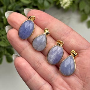 May include: Four pendants featuring smooth, polished, light purple stones set in gold-toned metal frames. Two pendants are oval-shaped, and two are teardrop-shaped. Each piece includes a small gold-toned bail at the top for attachment to a chain.
