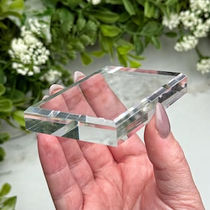 Puede incluir: Un prisma de cristal rectangular transparente con bordes biselados. El cristal se sostiene en una mano, con un fondo borroso de follaje verde y flores blancas. El prisma es transparente, permitiendo el paso de la luz.