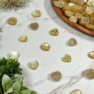 May include: A collection of heart-shaped sunstone crystals in various shades of yellow and gold. The crystals are arranged on a white marble surface with green succulents in the background.