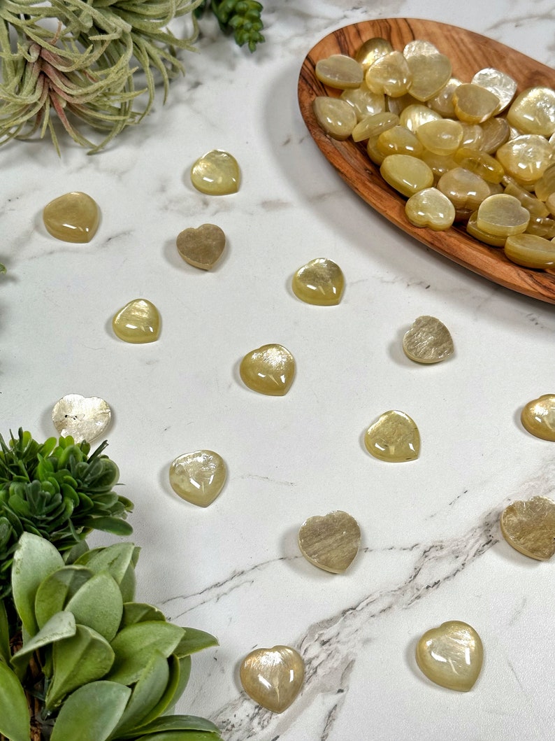 May include: A collection of heart-shaped sunstone crystals in various shades of yellow and gold. The crystals are arranged on a white marble surface with green succulents in the background.
