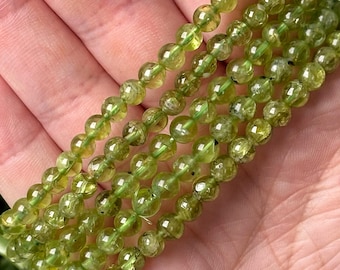 May include: Strands of round, translucent, light green peridot gemstone beads. The beads are strung together, showcasing their natural color and clarity. The beads are held in a hand, highlighting their size and shape.