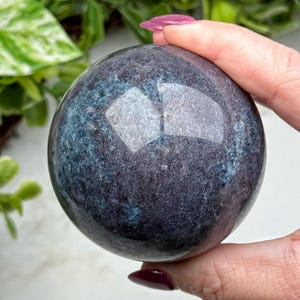 May include: A polished, spherical gemstone with a dark blue and purple color scheme. The surface reflects light, showcasing a glossy finish. The crystal ball is held in a hand, with green foliage in the background.