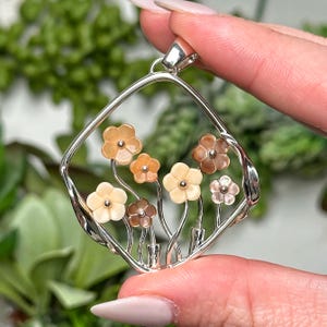 Peut inclure: Un pendentif argenté avec un cadre carré, présentant un motif floral. Le pendentif présente une collection de petites fleurs multicolores dans des tons crème, marron et orange. Les fleurs sont attachées à de fines tiges argentées.