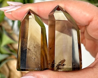 May include: Two smoky quartz crystal points, one with a darker brown hue and the other with a lighter brown hue, are held in a hand. The crystals are clear and have a slight shimmer.