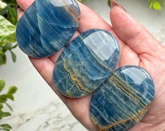 May include: Three polished, oval-shaped blue calcite stones with golden-brown veining are held in a hand. The stones have a glossy, reflective surface. The background includes green foliage and a white marble surface.