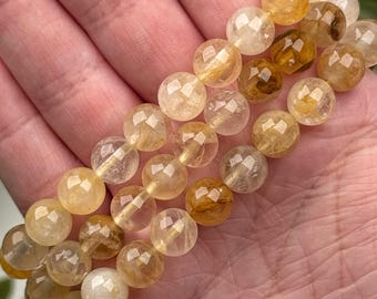 May include: A bracelet composed of faceted, round, translucent golden-yellow beads. The beads exhibit varying shades of amber, honey, and clear quartz, featuring internal inclusions and natural patterns that catch the light across their surfaces.