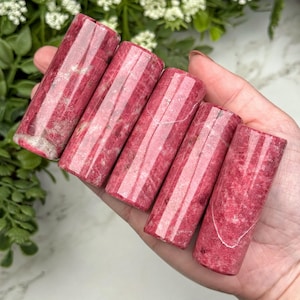 May include: Five cylindrical polished crystals in varying shades of deep magenta and pink. Each stone features a smooth, glossy surface with natural internal mineral inclusions and vertical striations, arranged in a row across the palm.