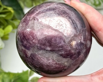 May include: A polished, spherical gemstone in shades of purple and white. The orb is held in a hand, showcasing its smooth, reflective surface. The stone's interior features a marbled pattern, creating visual depth and interest. The background includes green foliage.