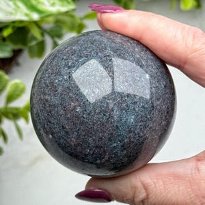May include: A dark, speckled sphere with reflective facets. The polished surface of the orb displays a mix of dark gray, blue, and green hues. The sphere is held in a hand, with a blurred green plant in the background.
