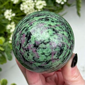 Può includere: Una pietra preziosa sferica lucidata caratterizzata da un mix vibrante di zoisite verde chiaro screziata, inclusioni di rubino magenta intenso e macchie minerali di orneblenda nera, con una superficie liscia e arrotondata.