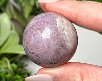 May include: A polished, spherical gemstone in shades of lavender and white. The orb is held between fingers, showcasing its smooth, reflective surface. The stone's color variations create a marbled effect, highlighting its natural beauty. This item is a decorative crystal.