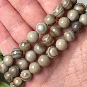 Puede incluir: Primer plano de un collar de cuentas redondas y pulidas. Las cuentas son principalmente beige con patrones arremolinados de marrón y blanco. Las cuentas se sostienen en una mano, mostrando su textura suave y variaciones naturales. Las cuentas tienen aproximadamente 1 cm de diámetro.