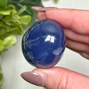 May include: A smooth, oval-shaped, blue crystal stone. The polished surface reflects light, creating bright highlights. The stone is held between fingers, with green foliage in the background. This crystal is a decorative item.