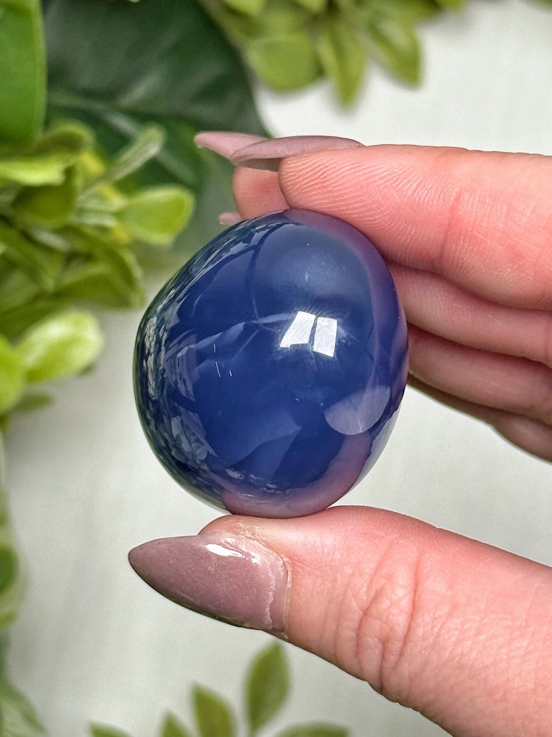 May include: A smooth, oval-shaped, blue crystal stone. The polished surface reflects light, creating bright highlights. The stone is held between fingers, with green foliage in the background. This crystal is a decorative item.