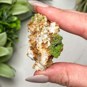 May include: A raw mineral specimen featuring a mix of colors and textures. The piece displays a base of white and tan, with clusters of vibrant green crystals. The rough, natural form of the stone is visible.