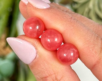 Peut inclure: Trois perles rondes de couleur rose, probablement en quartz rose, sont tenues dans une main. Les perles sont lisses et polies.