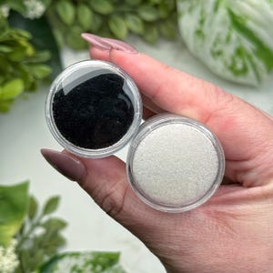 May include: Two small, clear, round containers held in a hand. One container is filled with black powder, the other with white powder. The background features green foliage.