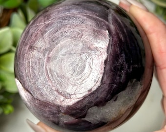 May include: A polished, spherical gemstone with swirling patterns in shades of purple, lavender, and white. The orb is held in a hand, showcasing its smooth, reflective surface. The stone's unique veining creates a captivating visual texture.