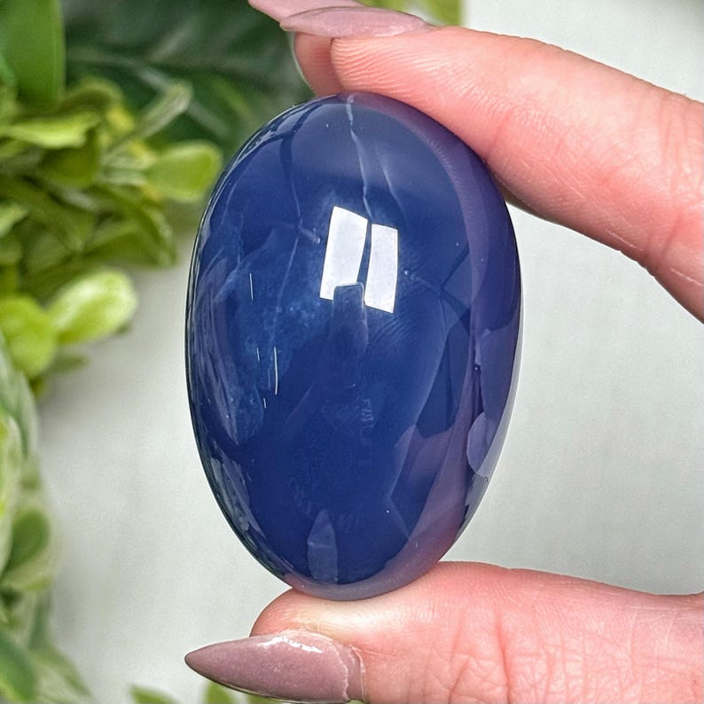 May include: A smooth, oval-shaped, deep blue gemstone is held between fingers. The polished stone reflects light, creating bright white highlights. The background features green foliage. The gemstone's shape and color suggest it could be used for jewelry or decorative purposes.