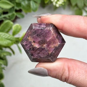 May include: A hexagonal-shaped gemstone with a deep magenta and dark red color palette. The stone features a polished surface with visible natural inclusions, dark mineral flecks, and light-colored veining throughout its crystalline structure.