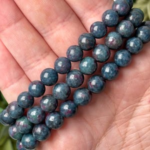 May include: Close-up of three strands of blue-green gemstone beads. Each bead is round and has flecks of red and purple. The beads are smooth and shiny, reflecting light. The beads are held in a hand.