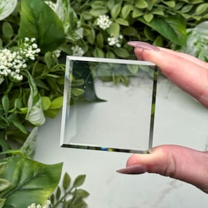 以下が含まれることがあります： 面取りされたエッジを持つ透明な正方形のガラスブロック。 ガラスは、緑の葉と白い花の背景に対して保持されています。 ガラスの端は光を反射し、微妙な虹色の効果を生み出します。