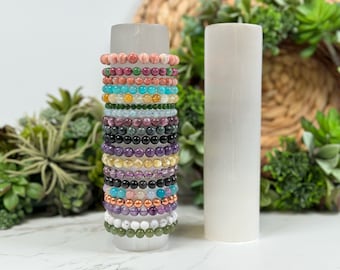 May include: A stack of colorful gemstone bracelets on a white selenite charging plate. The bracelets are made of various stones, including amethyst, rose quartz, and turquoise.