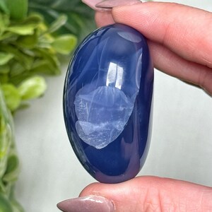 May include: A smooth, oval-shaped, polished blue gemstone. The stone has a deep, saturated blue color with lighter, translucent areas. The surface reflects light, highlighting its glossy finish. The gemstone is held between fingers, showcasing its size and shape.