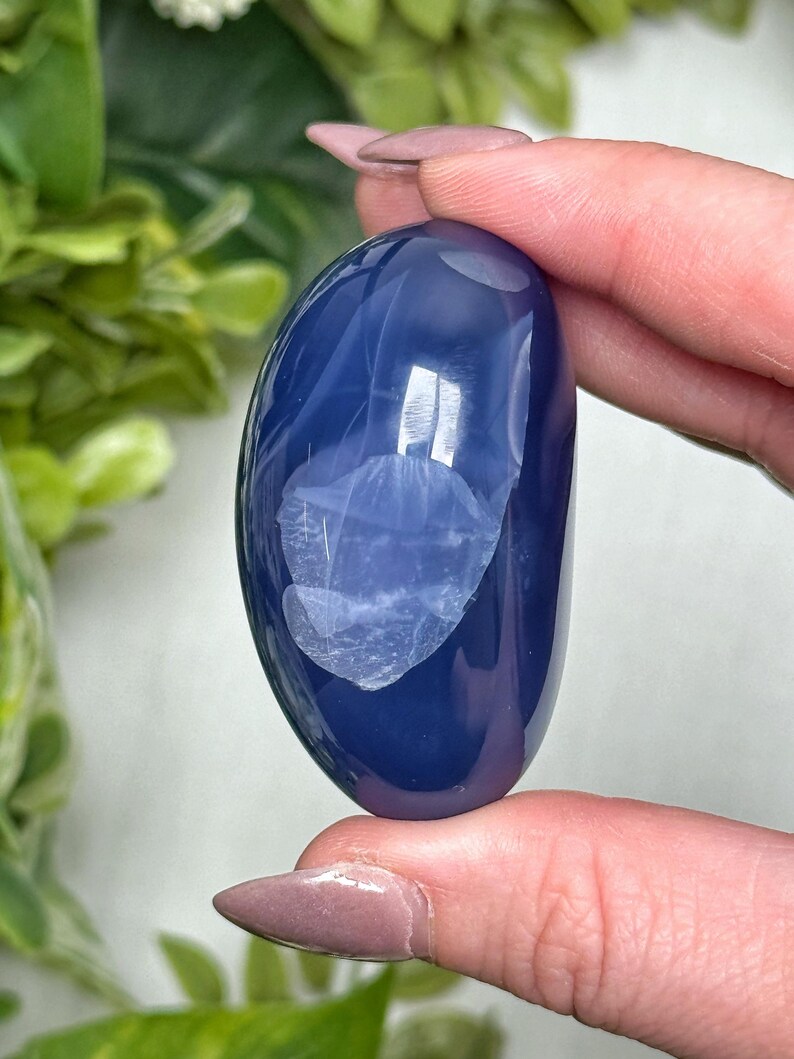May include: A smooth, oval-shaped, polished blue gemstone. The stone has a deep, saturated blue color with lighter, translucent areas. The surface reflects light, highlighting its glossy finish. The gemstone is held between fingers, showcasing its size and shape.